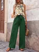 Woman wearing a floral top and green pants standing against a textured wall.