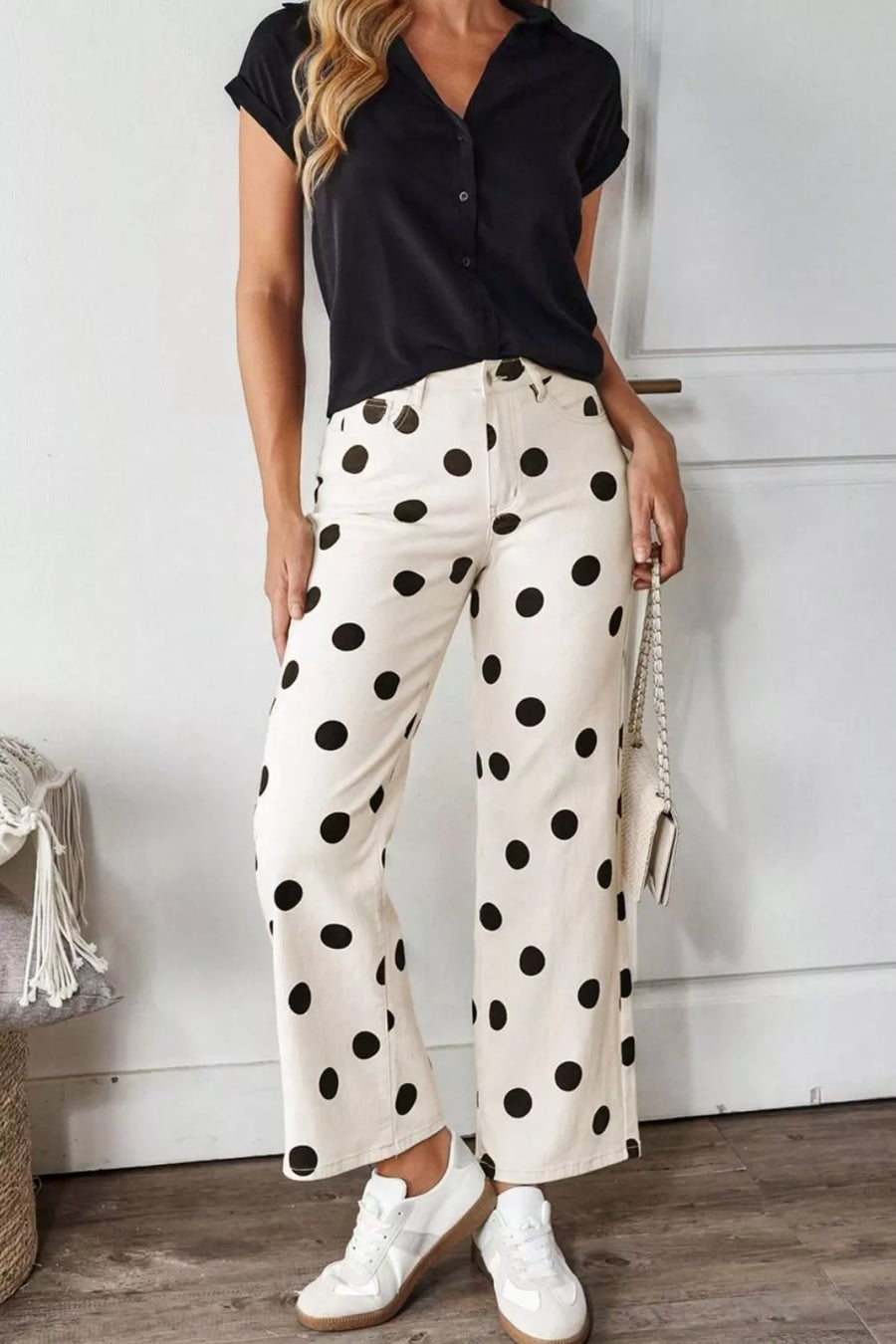 Woman wearing a black sleeveless top and white pants with black polka dots, standing indoors.