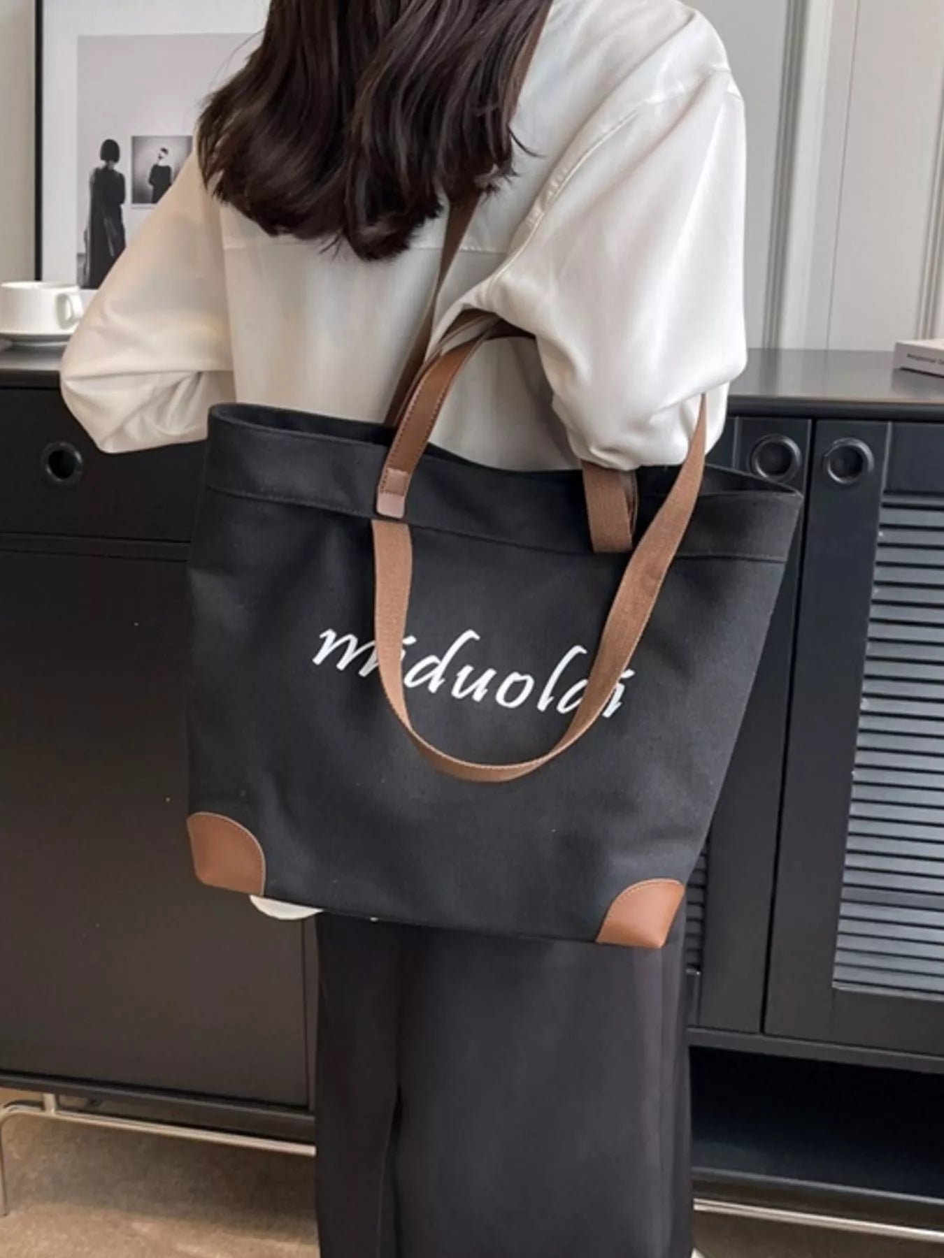 Person holding a black tote bag with 'miduola' branding in an indoor setting.