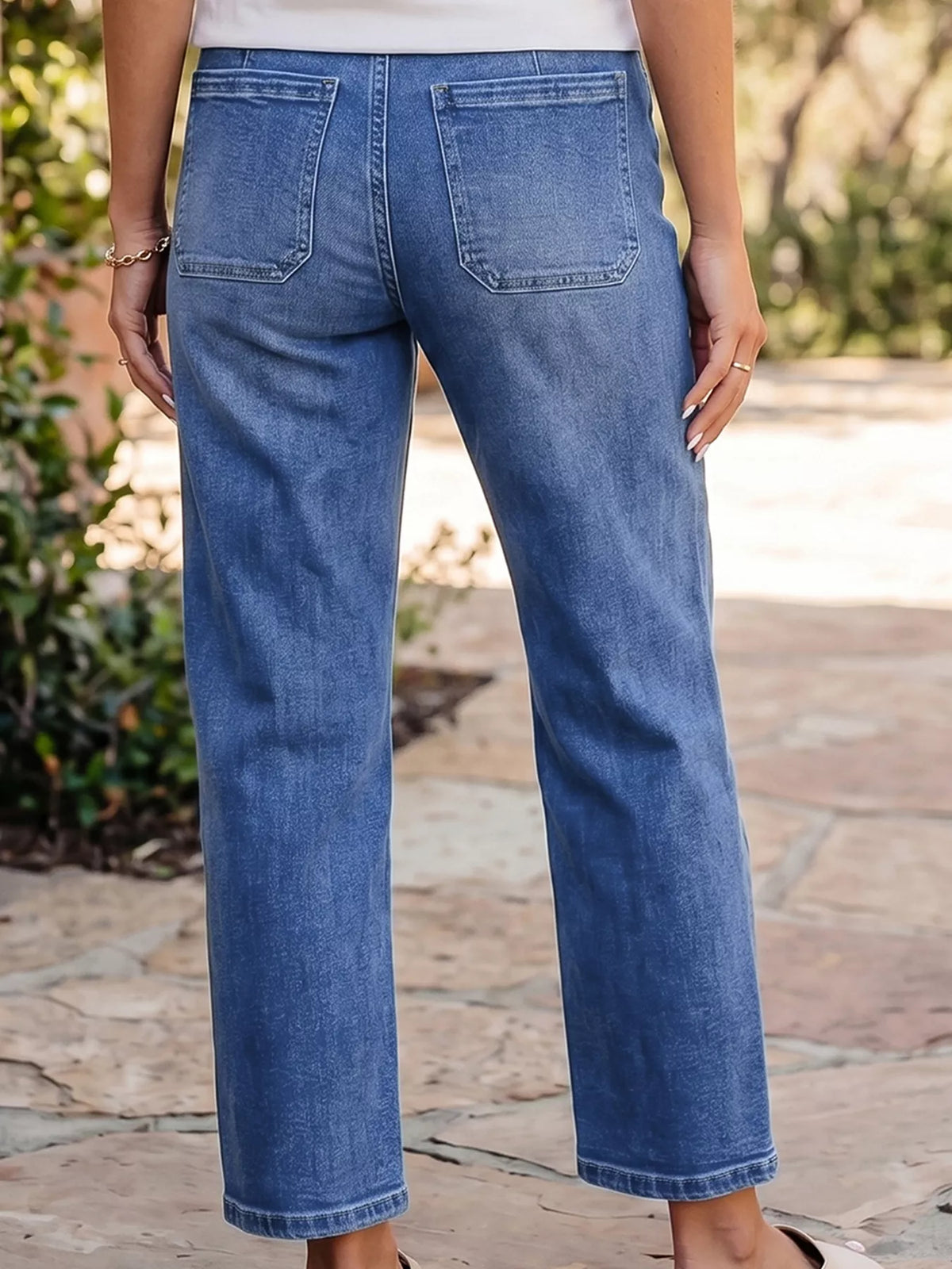A person viewed from the back wearing medium-wash blue cropped jeans with a straight-leg fit, standing outdoors on a stone walkway with greenery in the background.