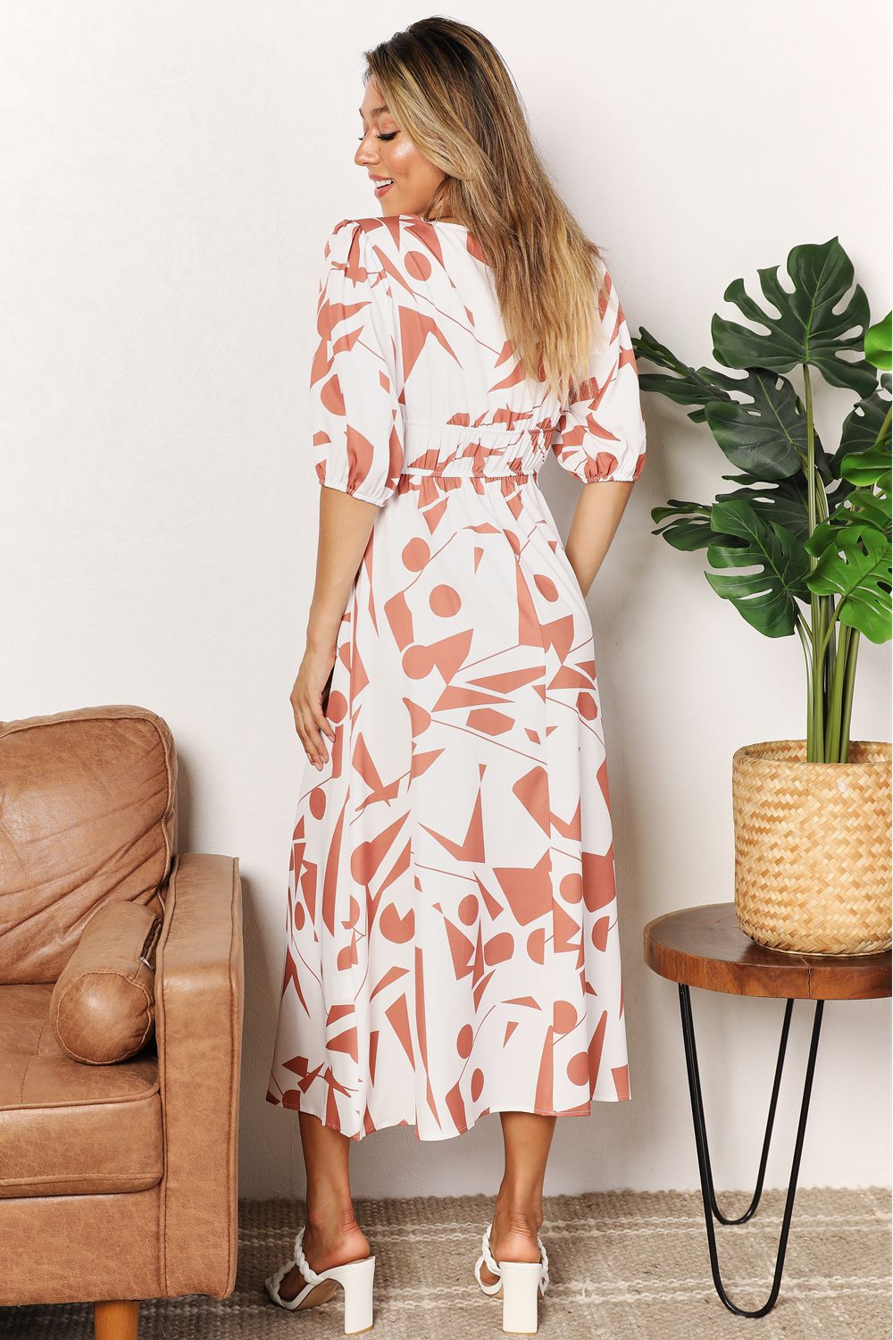 Woman in a floral dress standing in a room with a brown leather chair and a plant on a wooden table.