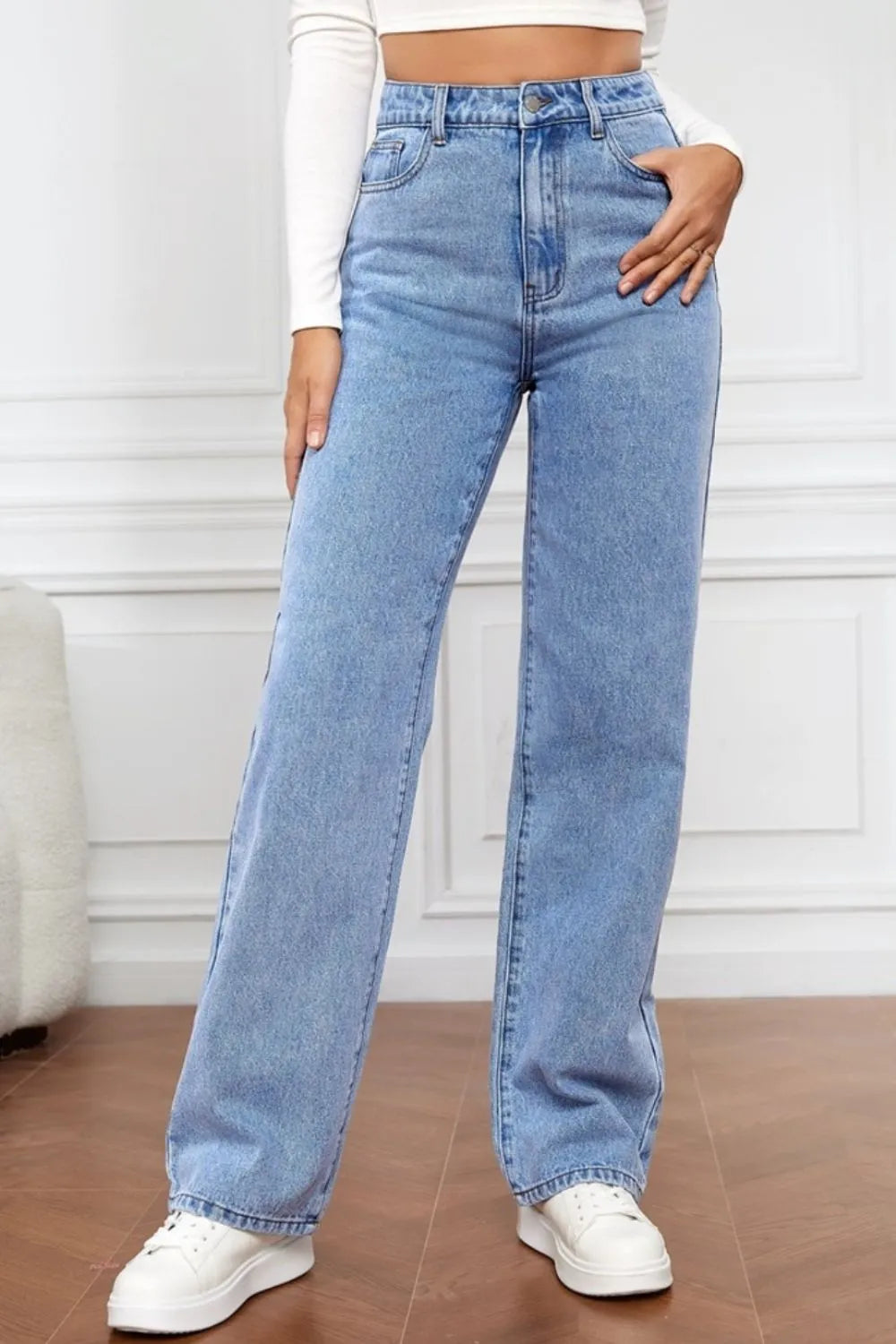 Person wearing light blue jeans and white sneakers on a wooden floor.