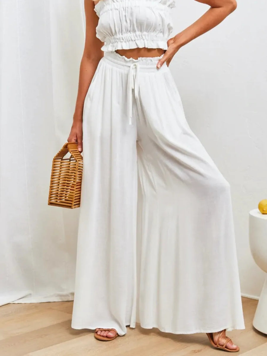 Person wearing a white outfit with wide-leg pants and a woven handbag indoors.