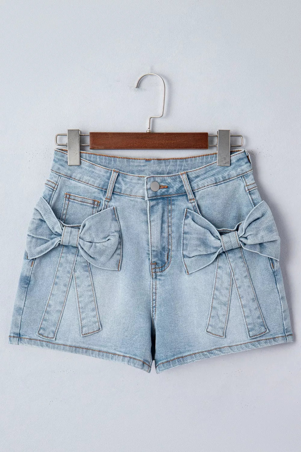 Light blue denim shorts with bow details on a hanger against a white background