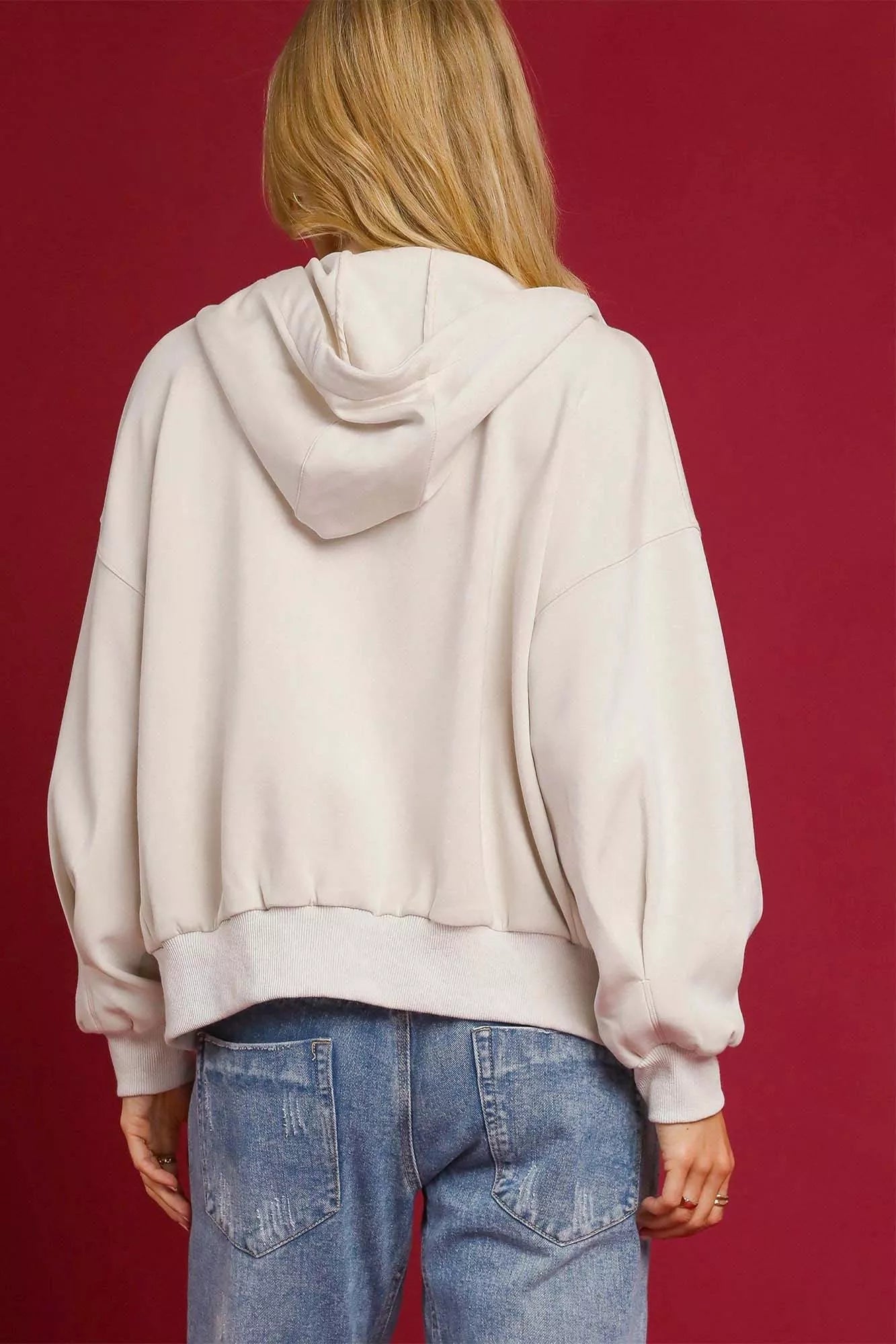 Person wearing a beige hoodie and blue jeans against a red background