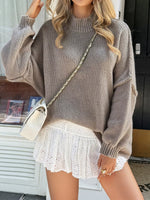 Woman wearing a brown sweater and white lace skirt with a beige handbag.