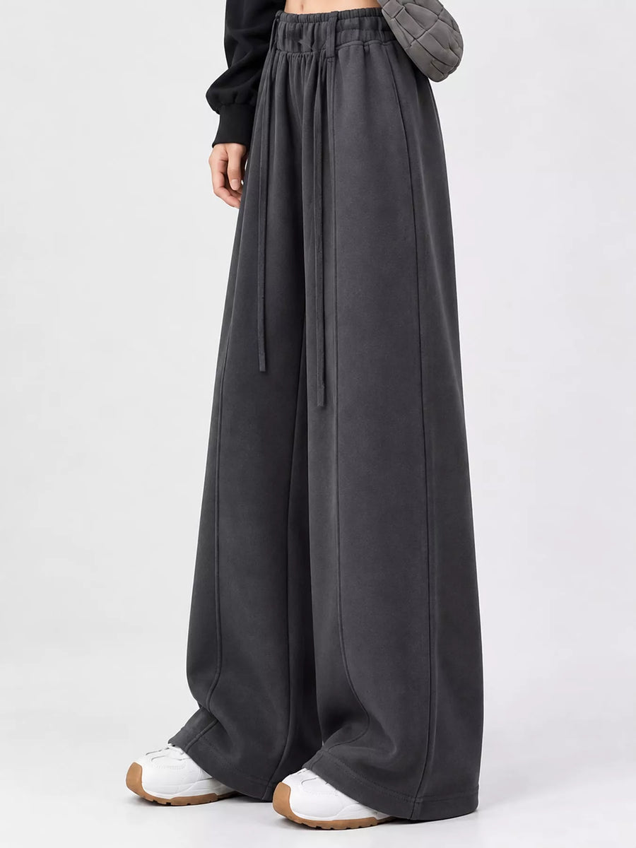 A person wearing very wide-leg dark charcoal gray sweatpants with a drawstring waist, styled with white sneakers against a light gray studio background.