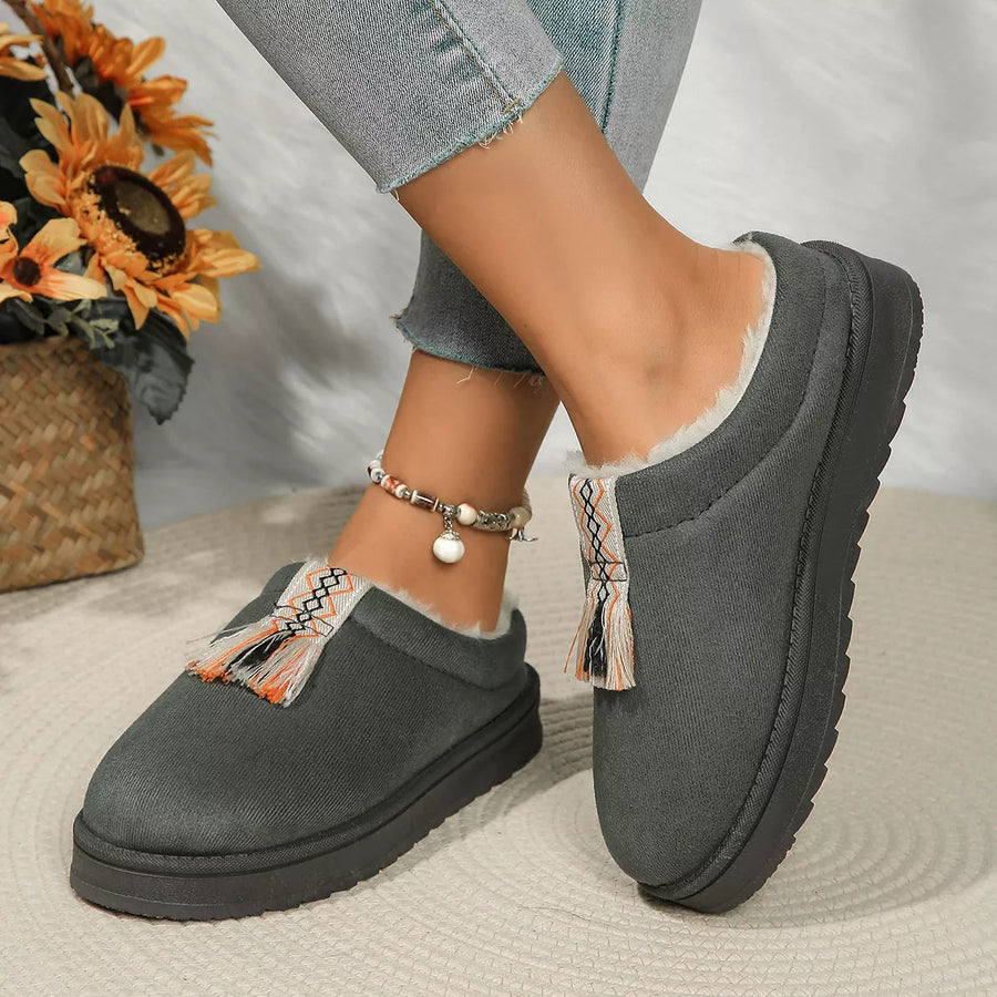 Gray slippers with tassels worn by a person, with a sunflower arrangement in the background.