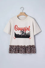 Beige t-shirt with 'Cowgirl' graphic and leopard print hem on a hanger against a light gray background