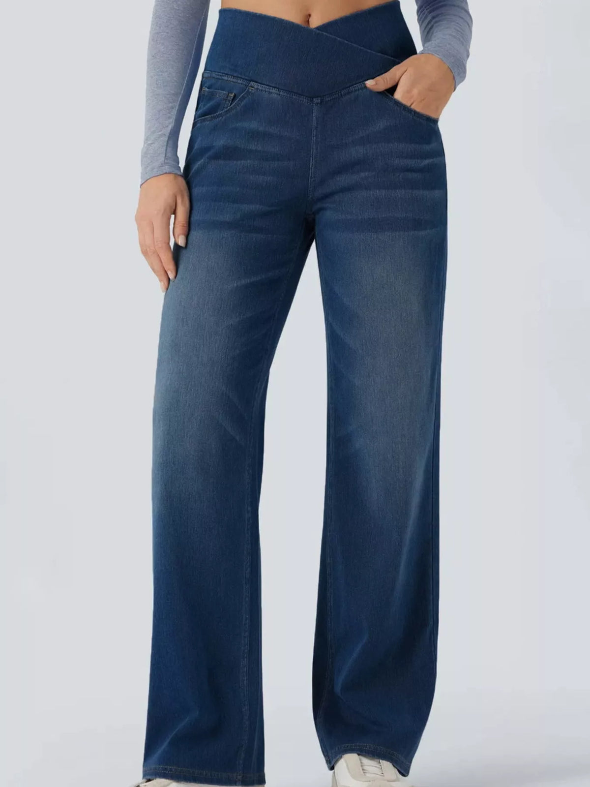 A person wearing dark blue wide-leg denim jeans with a crossover waistband, paired with a gray long-sleeve crop top and white sneakers, shown against a light gray studio background.