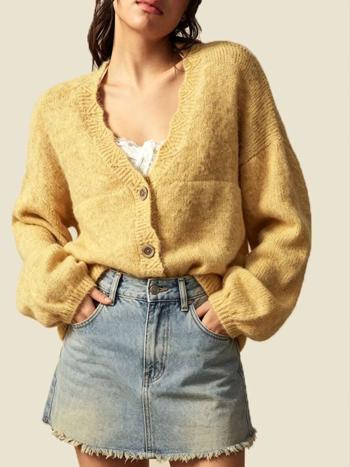 A woman wearing a fuzzy mustard yellow cardigan sweater with a V-neck and three buttons, paired with a light blue denim mini skirt, against a solid beige background.