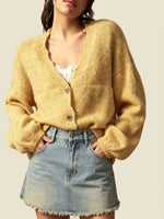 A woman wearing a fuzzy mustard yellow cardigan sweater with a V-neck and three buttons, paired with a light blue denim mini skirt, against a solid beige background.