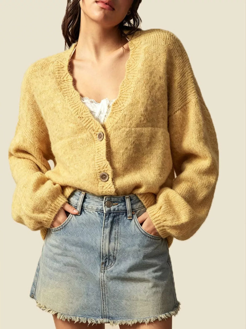 A woman wearing a fuzzy mustard yellow cardigan sweater with a V-neck and three buttons, paired with a light blue denim mini skirt, against a solid beige background.