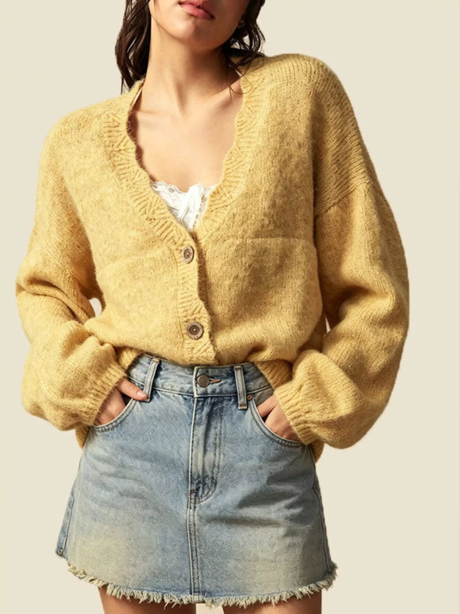 A woman wearing a fuzzy mustard yellow cardigan sweater with a V-neck and three buttons, paired with a light blue denim mini skirt, against a solid beige background.