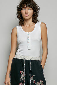 Woman wearing a white sleeveless top with colorful buttons and black pants with floral embroidery on a gray background