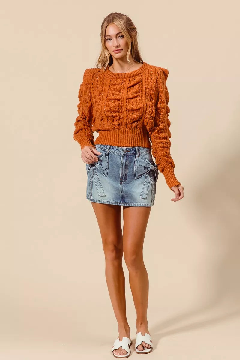 Woman wearing an orange cable knit sweater and denim skirt on a beige background