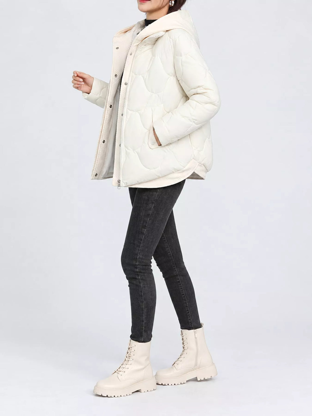 A model wearing a cream-colored quilted puffer jacket, black skinny jeans, and matching cream lace-up boots, shown from the waist up against a light gray studio background.