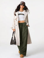 Woman wearing a white trench coat, green pants, and a cap on a white background
