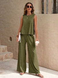 Woman wearing a green sleeveless top and wide-leg pants standing against a beige wall.