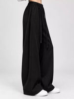 Black wide-leg pants with an elastic waistband and drawstring, shown on a model from the waist down wearing white sneakers against a light gray studio background.