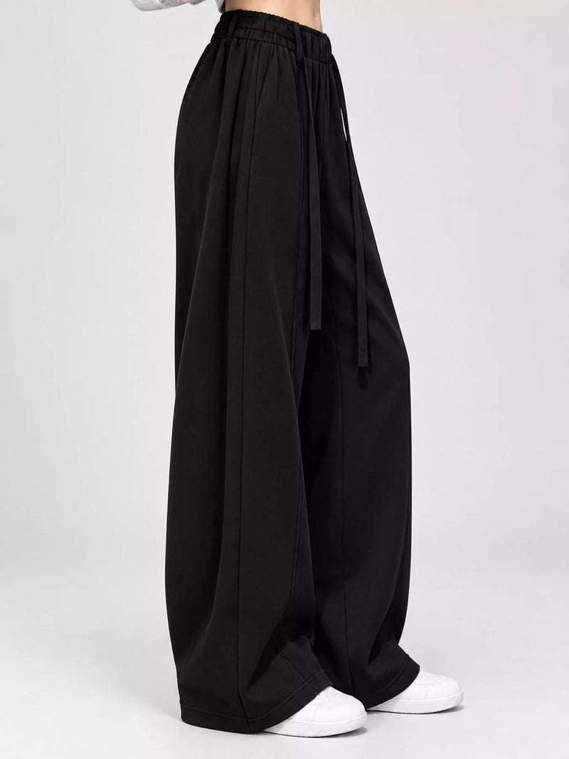 Black wide-leg pants with an elastic waistband and drawstring, shown on a model from the waist down wearing white sneakers against a light gray studio background.