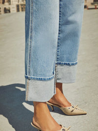 Close-up of rolled-up light blue jeans and beige mules on a blurred street background