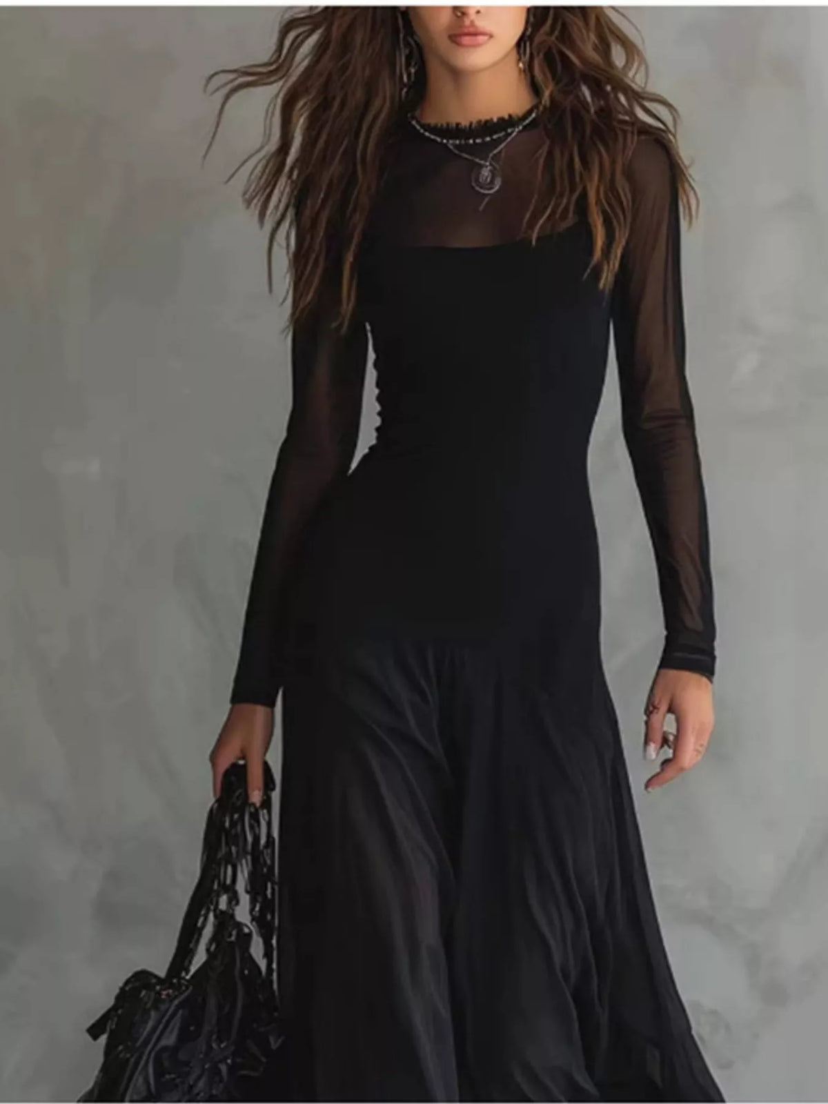 A woman wearing a black long-sleeve dress with sheer mesh sleeves and a high neckline, holding a black handbag against a textured gray studio background.