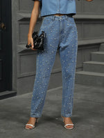 Person wearing blue jeans with pearl embellishments standing on a gray pavement.