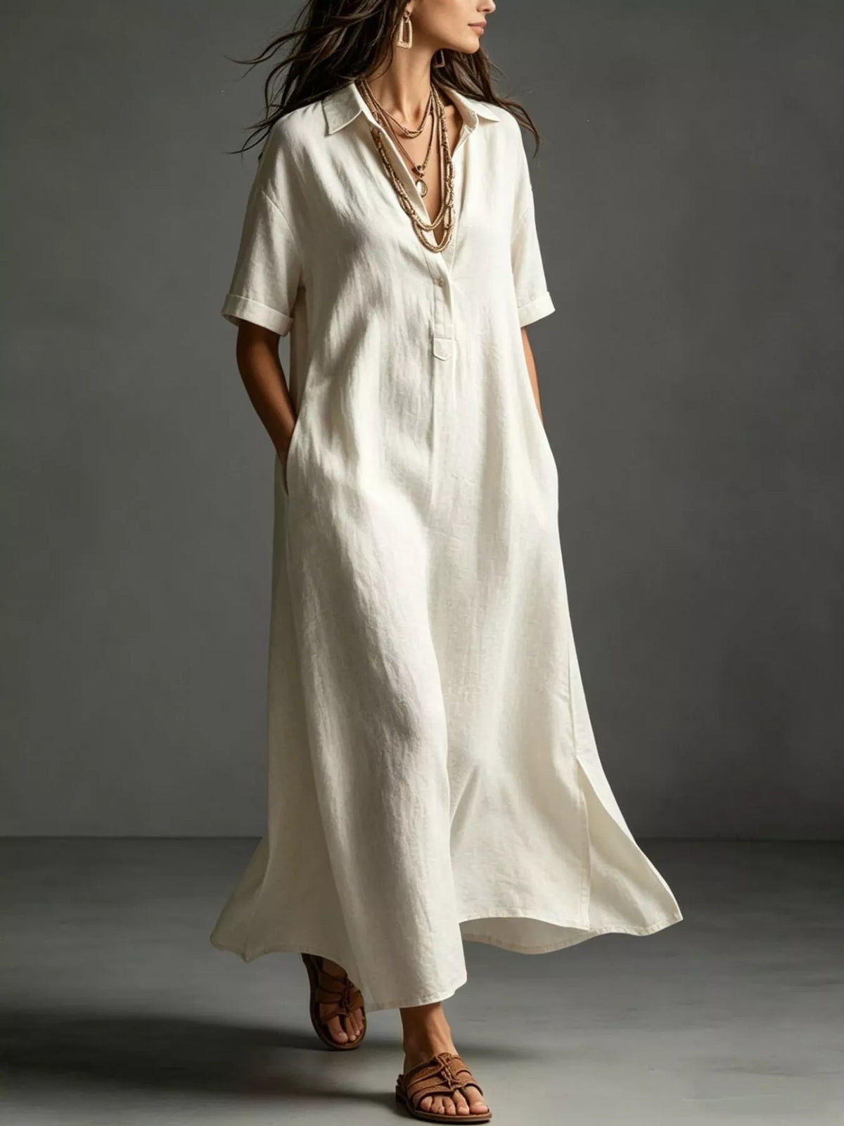A woman wearing a loose, short-sleeve white linen shirt dress with pockets, styled with layered gold necklaces and brown leather sandals, set against a dark gray studio background.