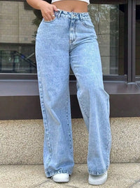 Person wearing light blue jeans standing on a sidewalk.