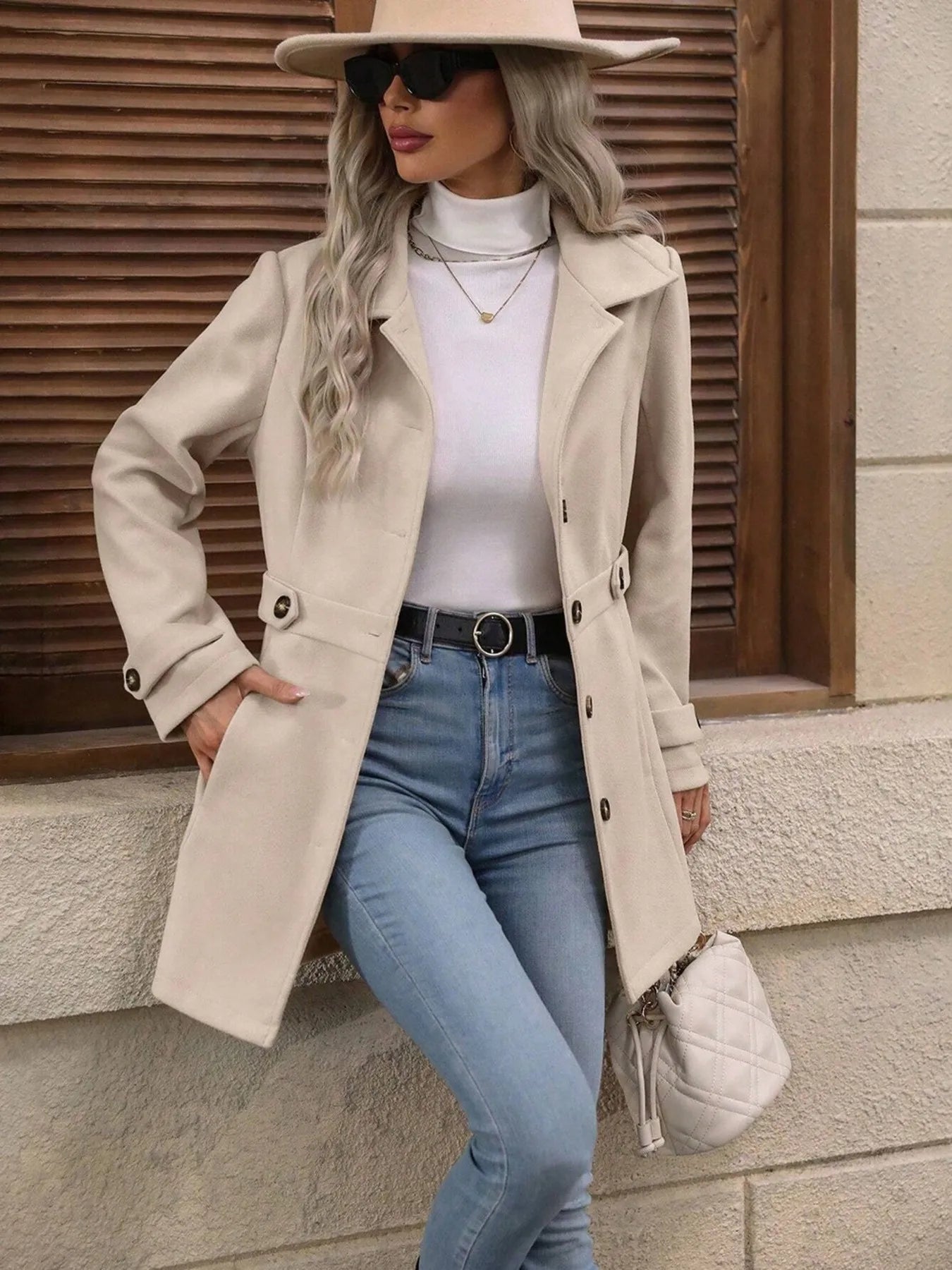 Woman wearing a beige trench coat, white sweater, blue jeans, and a beige hat against a wooden wall.
