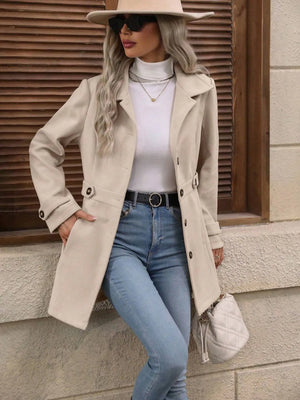 Woman wearing a beige trench coat, white sweater, blue jeans, and a beige hat against a wooden wall.
