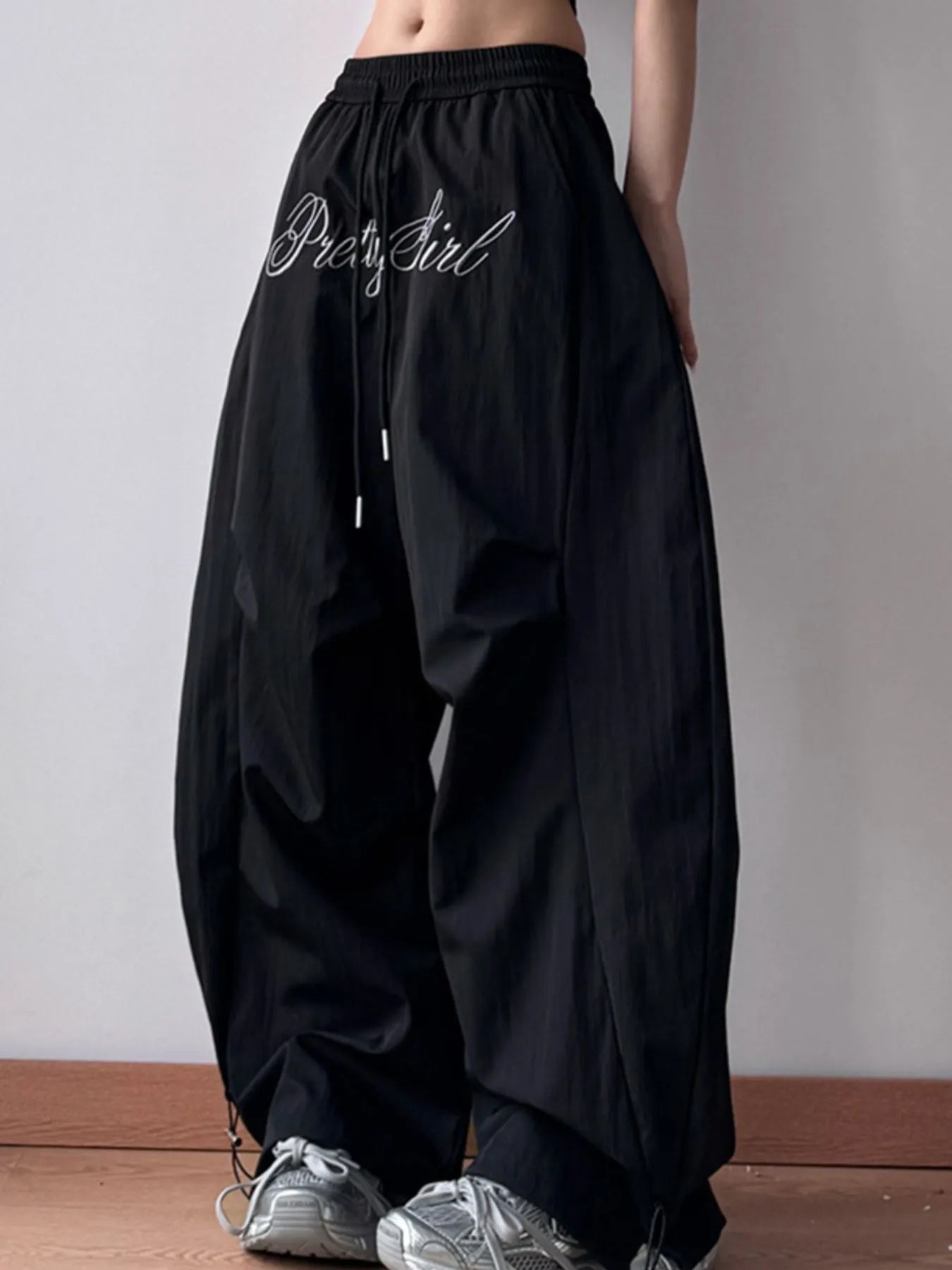 Black wide-leg pants with 'Pretty Girl' text worn by a person on a plain background