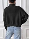 Person wearing a black sweater and light blue jeans against a white paneled wall.