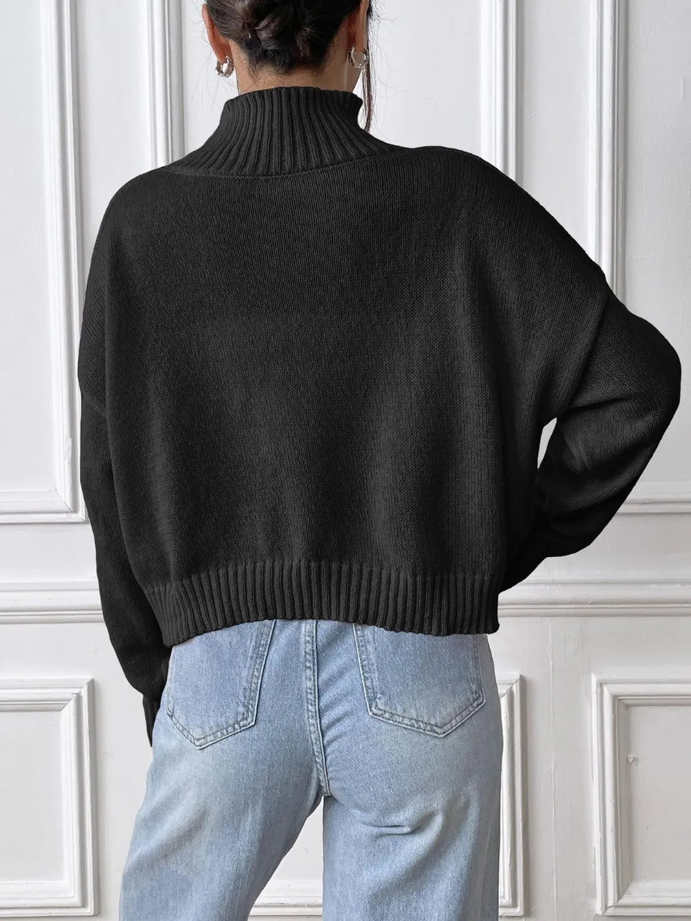 Person wearing a black sweater and light blue jeans against a white paneled wall.