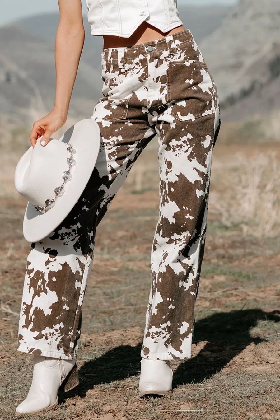 A person outdoors wearing brown and white mottled cow-print pants, a white cropped top, white cowboy boots, and holding a white cowboy hat with silver embellishments. The background shows a dry grassy field and hills.