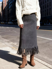 Person wearing a white cable knit sweater, gray fringed skirt, and brown boots on a city street.