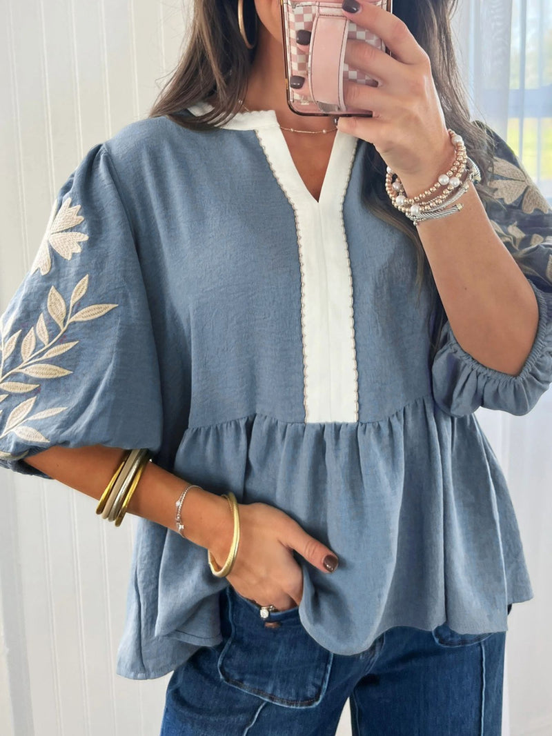 Woman wearing a blue blouse with floral embroidery and white trim, taking a mirror selfie.