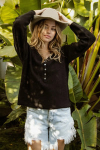 Woman wearing a black long-sleeve shirt and white shorts with a beige hat, standing among green plants.