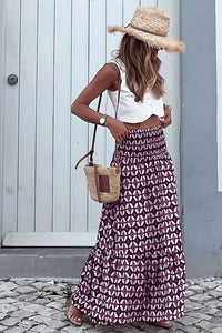 Woman wearing a white top and patterned skirt with a straw hat and bag, standing against a light blue wall.