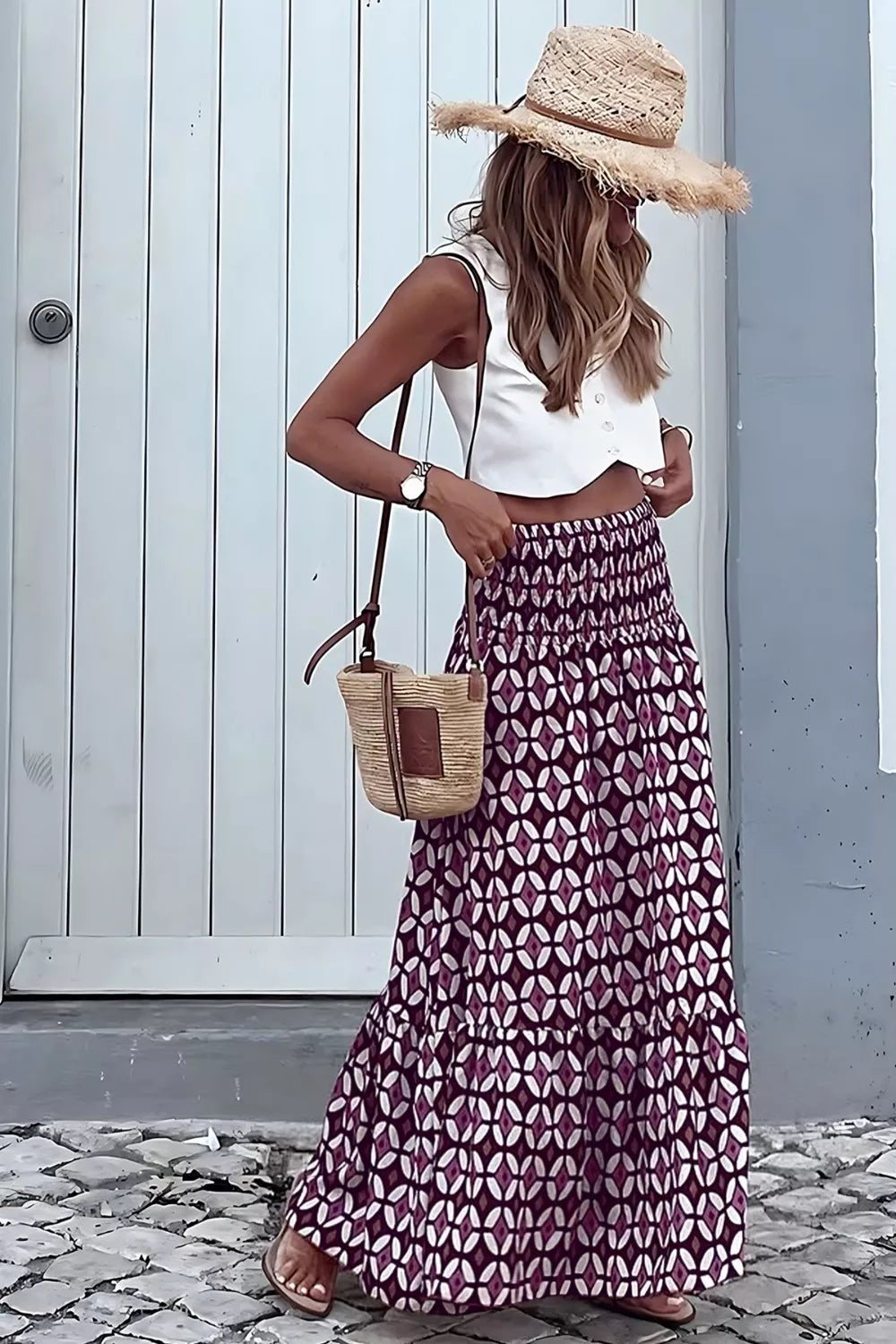 Woman wearing a white top and patterned skirt with a straw hat and bag, standing against a light blue wall.
