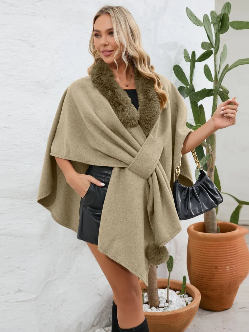 Woman wearing a beige poncho with fur trim, standing next to potted plants.