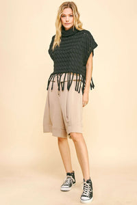 Woman wearing a dark green knitted poncho with fringe, beige shorts, and black high-top sneakers on a beige background.