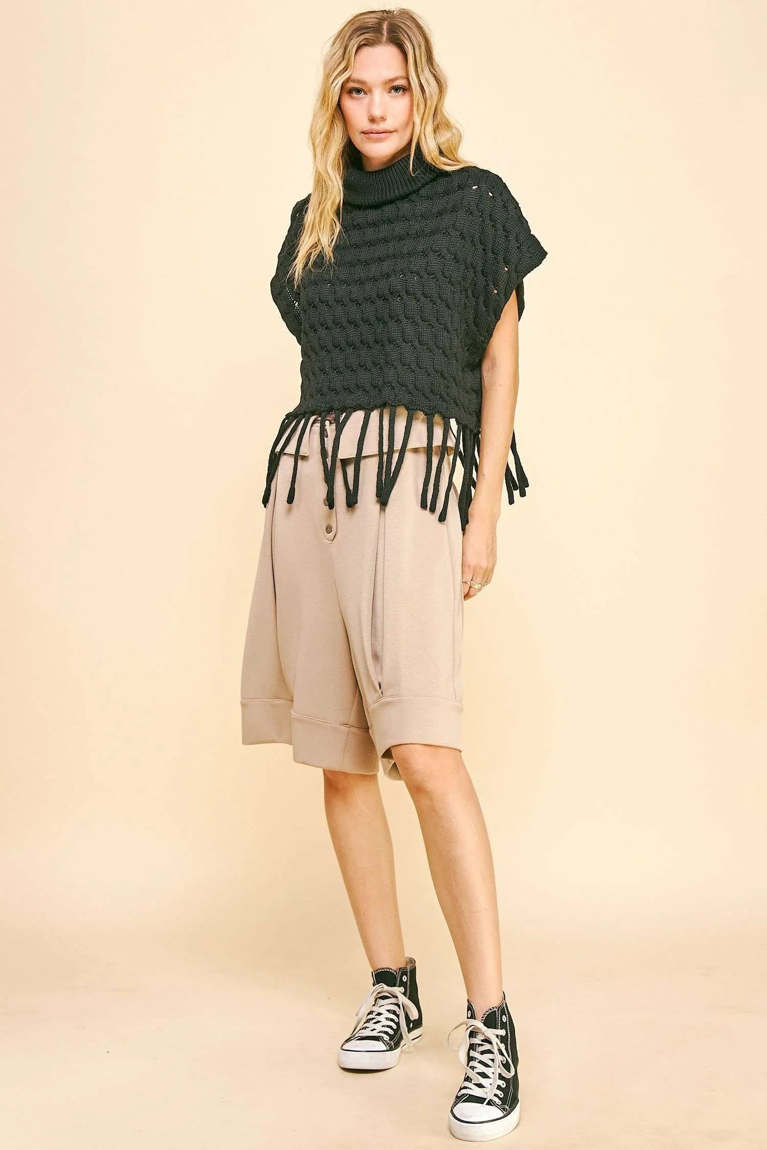 Woman wearing a dark green knitted poncho with fringe, beige shorts, and black high-top sneakers on a beige background.