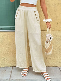 Person wearing cream-colored wide-leg pants with button details, holding a small bag against a beige wall.