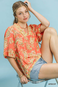 Woman wearing a patterned orange top and denim shorts sitting on a chair against a light blue background.