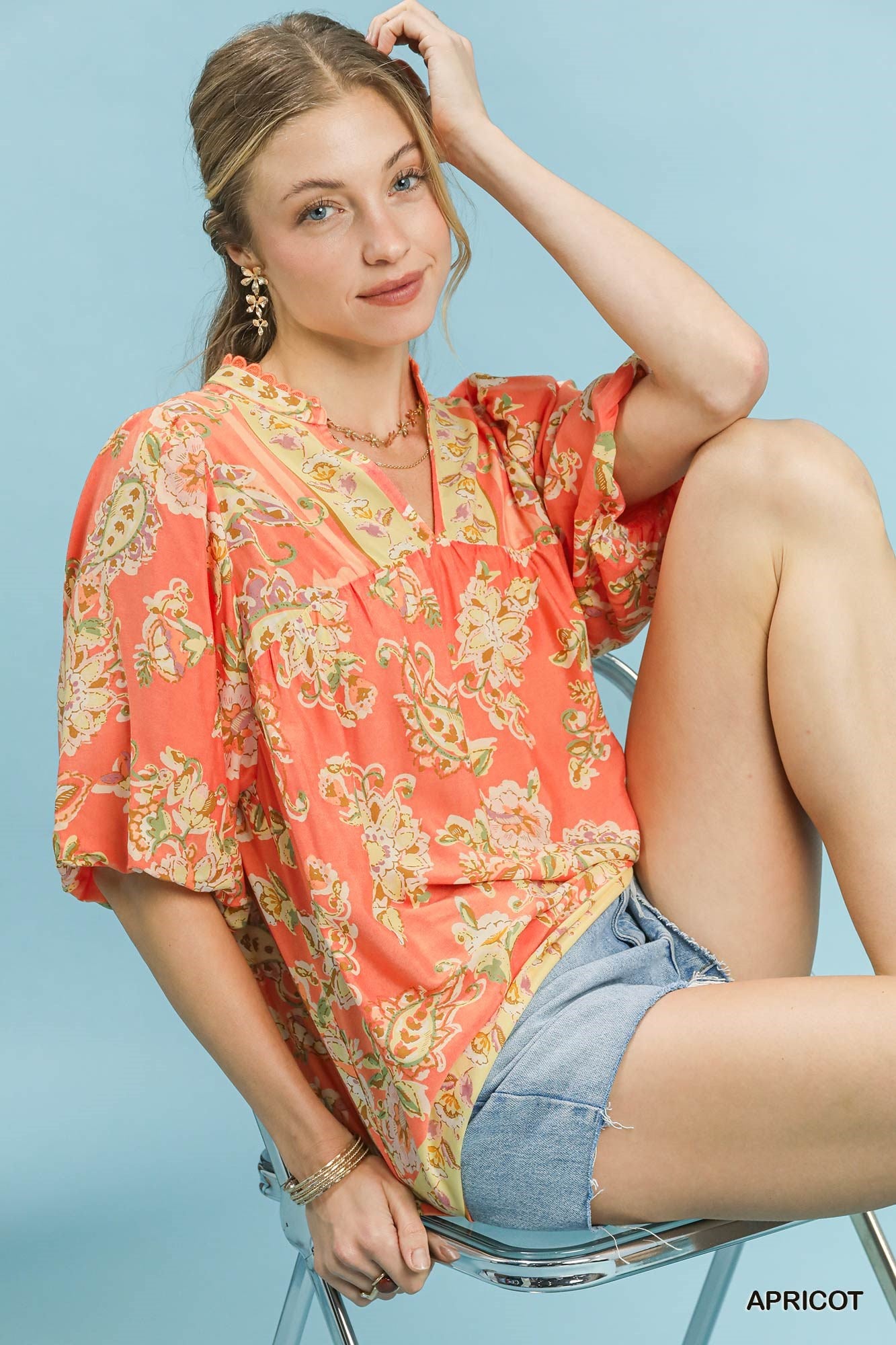 Woman wearing a patterned orange top and denim shorts sitting on a chair against a light blue background.