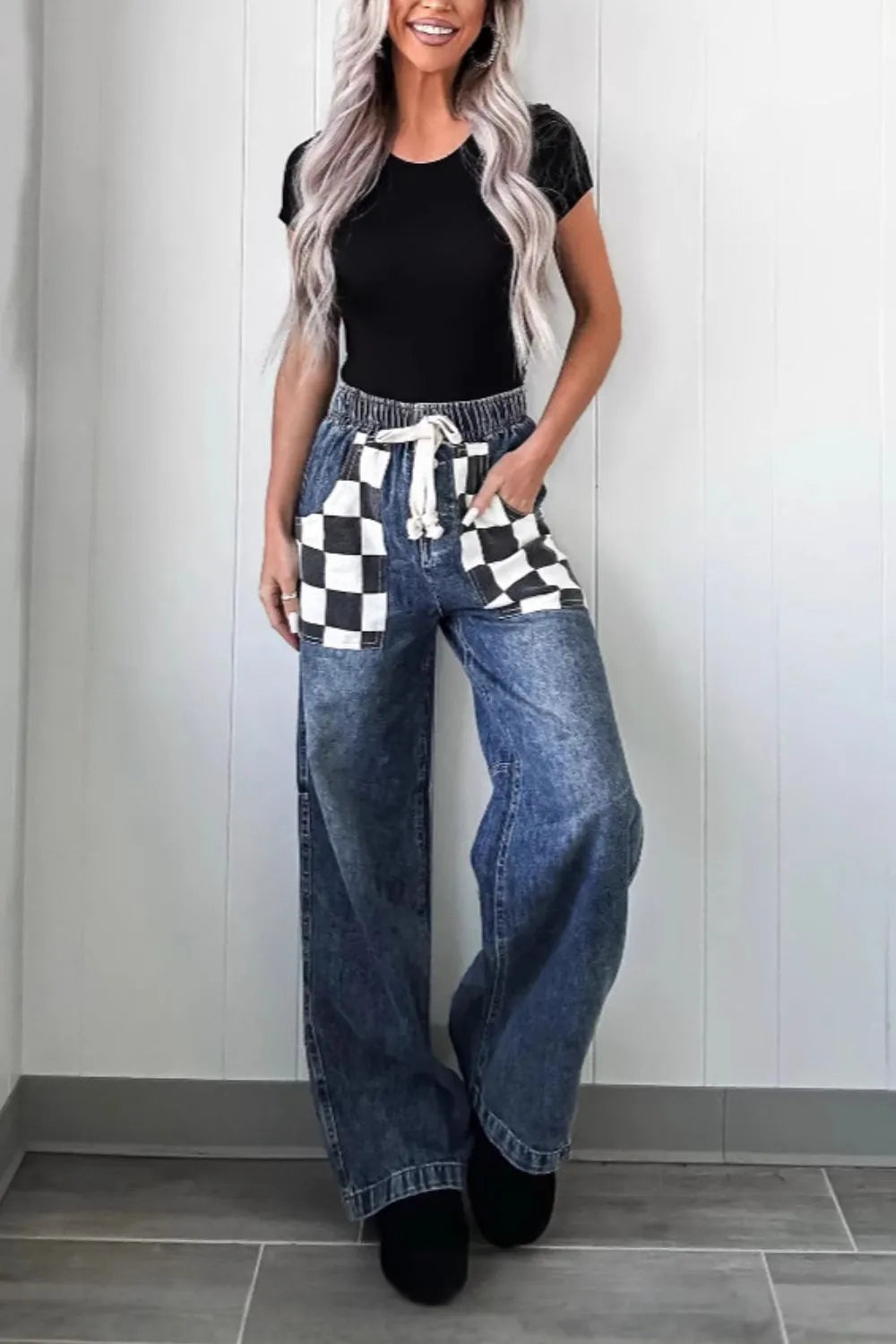 Woman wearing a black top, blue jeans, and a checkered apron against a white wall.