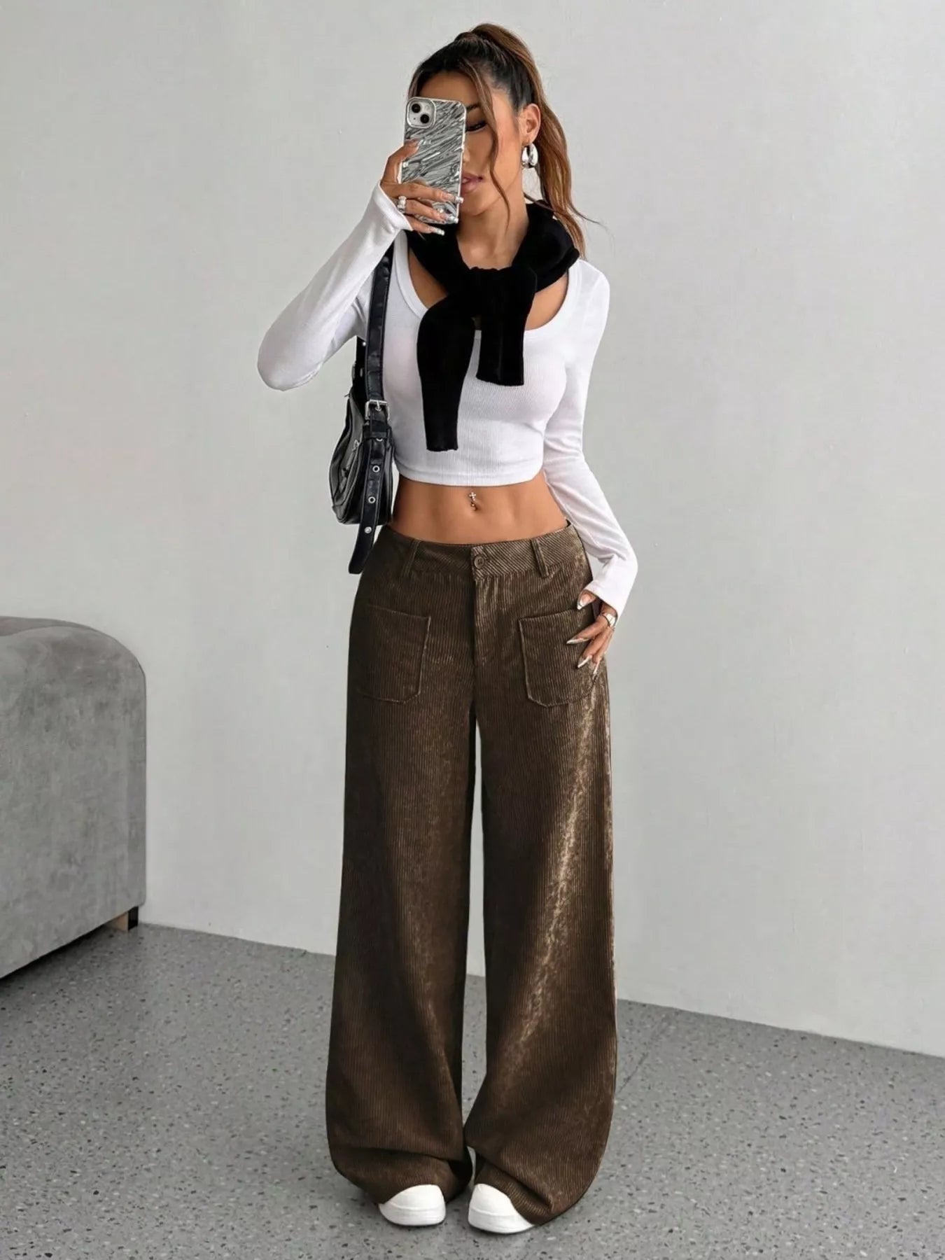 Person wearing a white long-sleeve top with a black scarf and brown wide-leg pants, standing against a plain background.