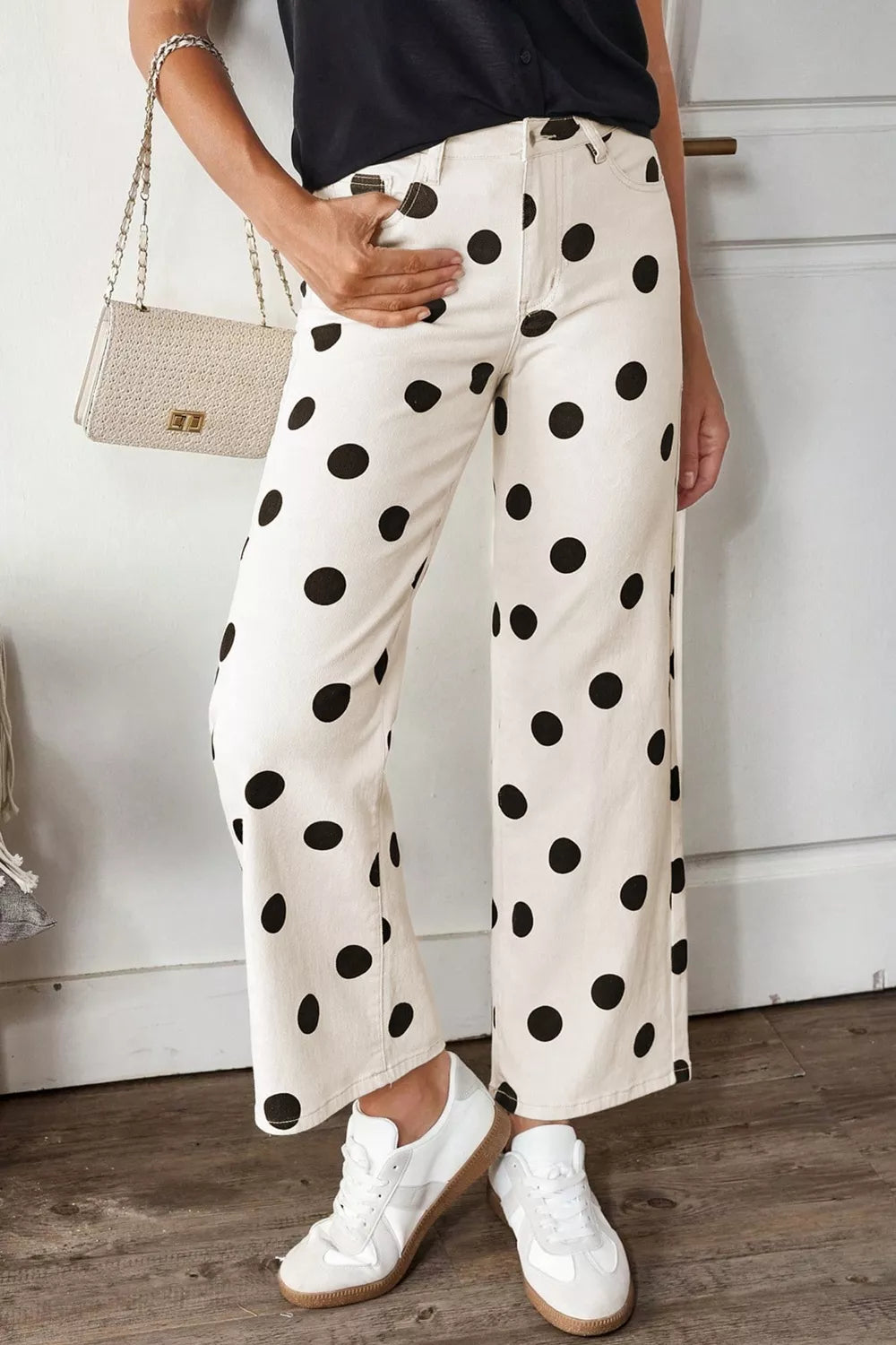 Person wearing white pants with black polka dots, white sneakers, and a black top, holding a beige handbag.